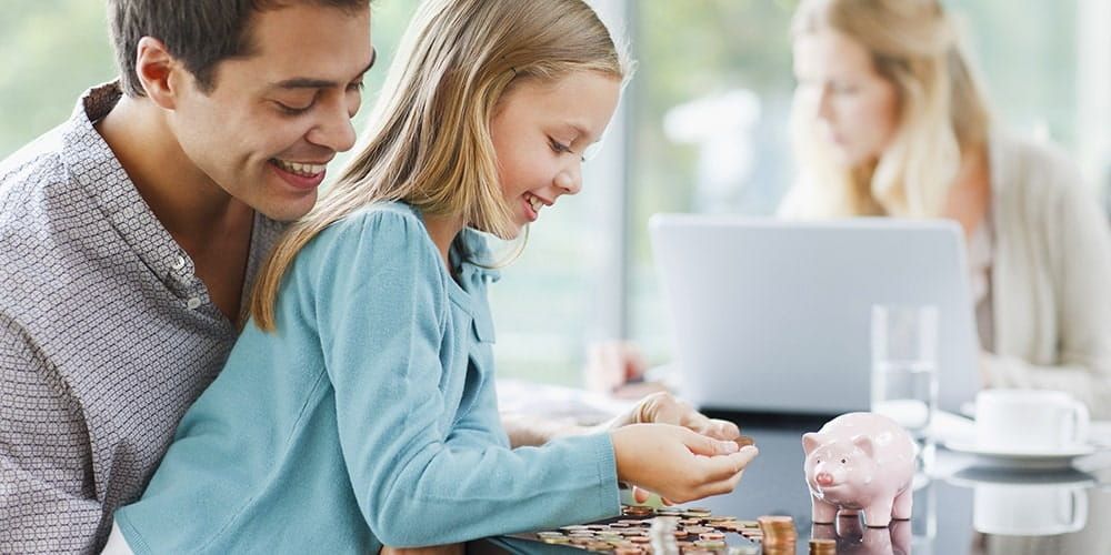 Father sits with young daughter on his knee as they smile together whilst putting coins into a pink piggy bank in front of them
