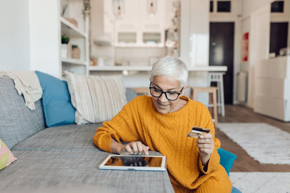 Woman in yellow sweater is sat on the floor of her living room leaning against her sofa. She is holding her bank card and entering it's details into her ipad