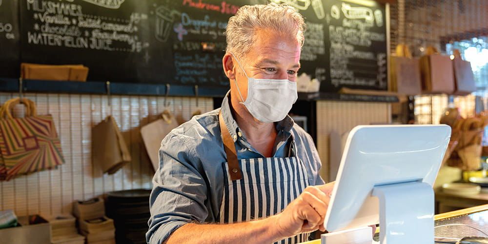 Older man with stripped apron working at a till in a cafe, wearing a surgical mask