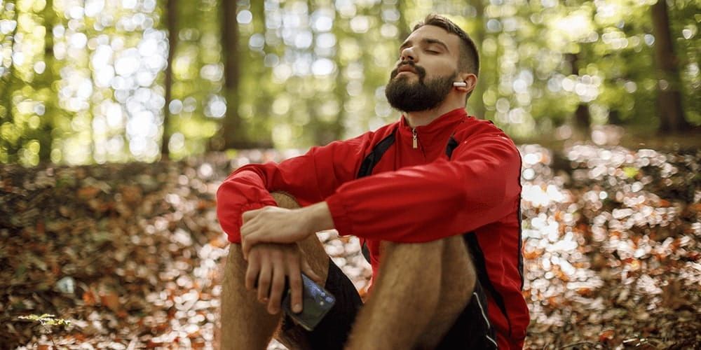 Man sits in running gear in the middle of a wooded area. He is holding his phone with earphones in, closing his eyes and trying to relax