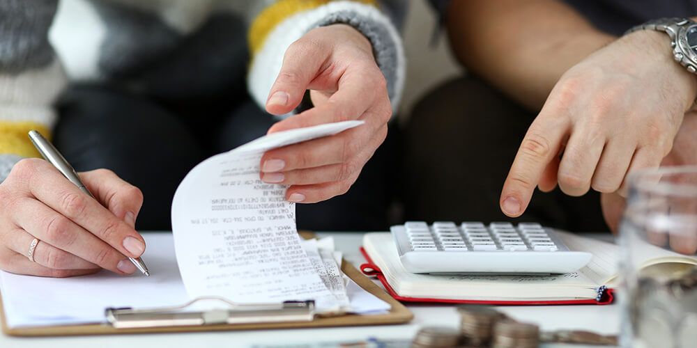 A close up shot of a persons hand holding a pen and looking at some receipts, while another person uses a calculator