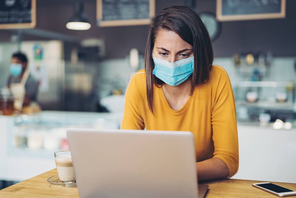Woman sat in cafe working at her laptop, wearing a surgical mask