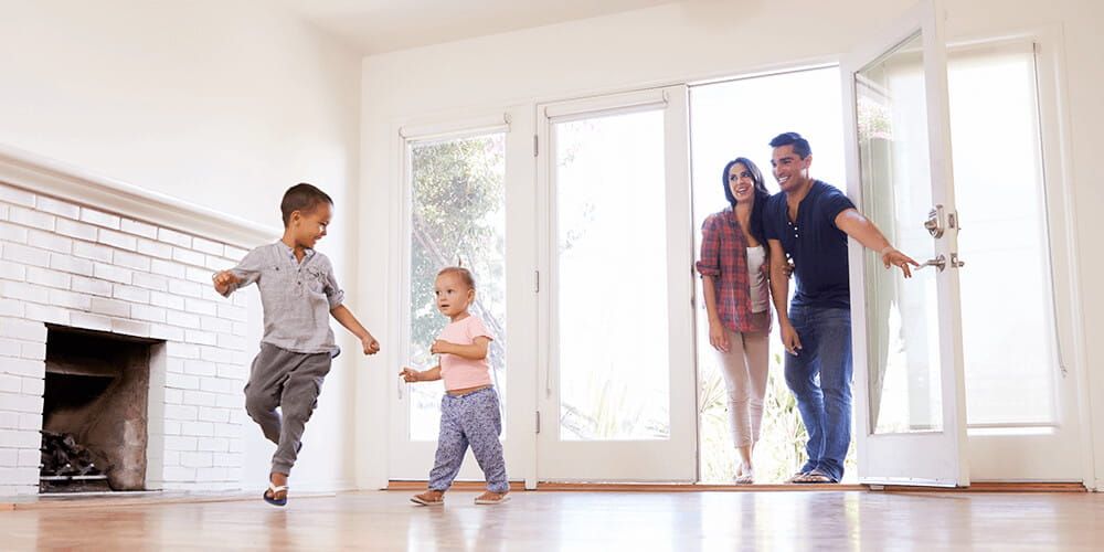 A family of four entering their new home together. The children run into the empty living room whilst the parents smile at them