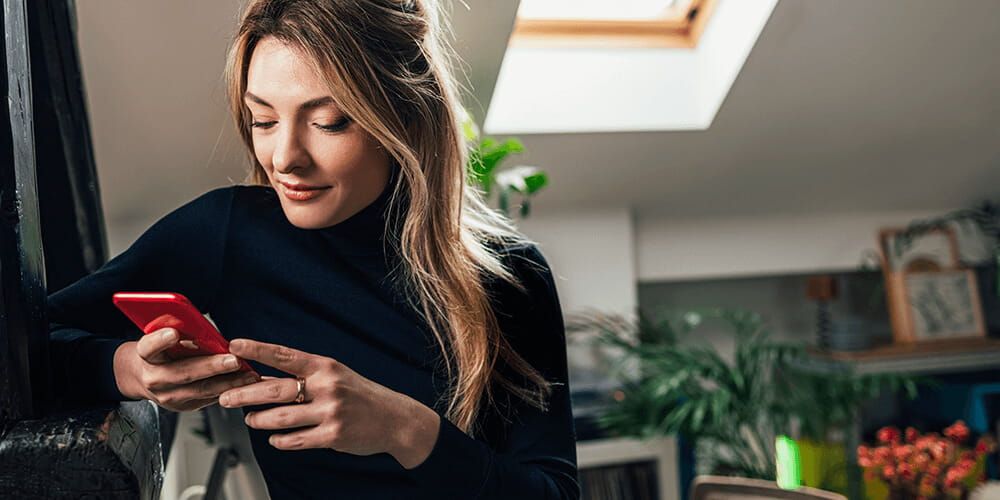 Woman stood in living room full of plants, looking down at her red phone with a happy face