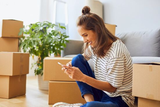 Young woman, wearing her hair in a bun, on her phone whilst surrounded by cardboard boxes after moving into her home