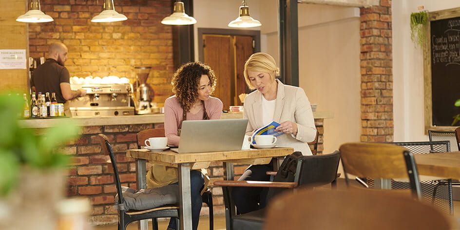 Two women sat togther in a coffee shop. One is on her laptop and the other is showing her pages in a brochure