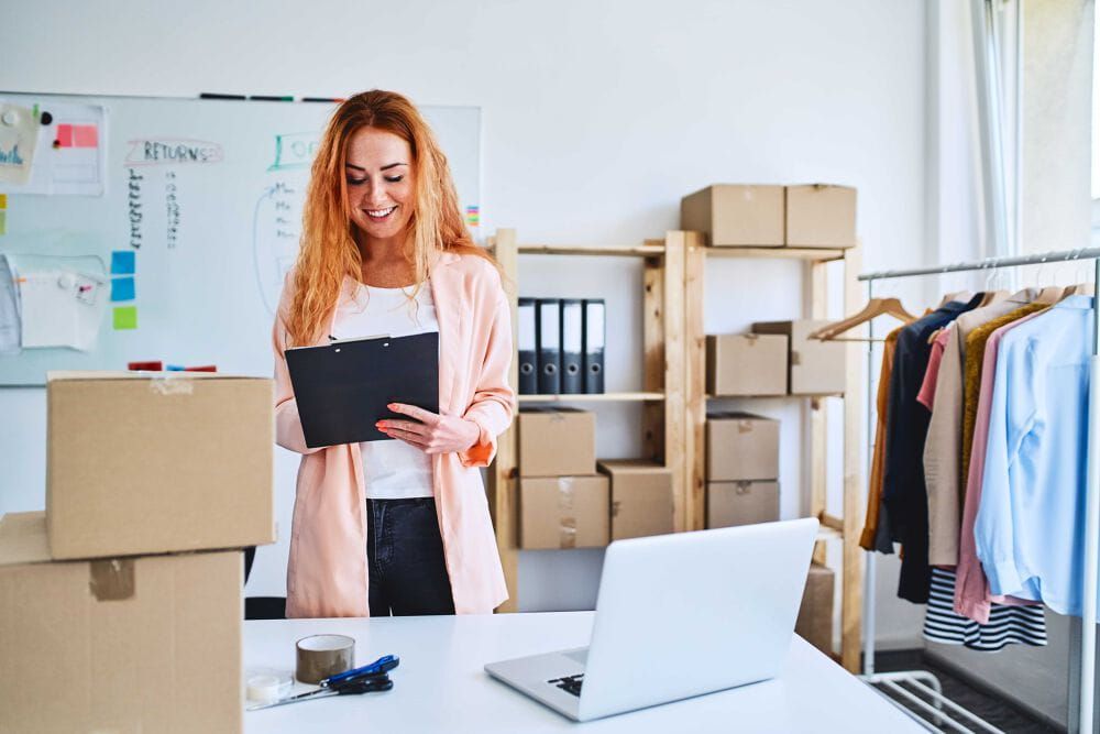 Woman standing in her fashion business office. She is holding a black clipboard and writing on it as she smiles. There is a large whiteboard behind her, many cardboard boxes around the office, a clothes rail and a laptop on her desk